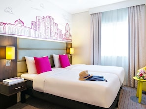 Una habitación de hotel con una cama grande, dos mesitas de noche, un cabecero con un mural de paisaje urbano, dos lámparas de pared y una ventana con cortinas.