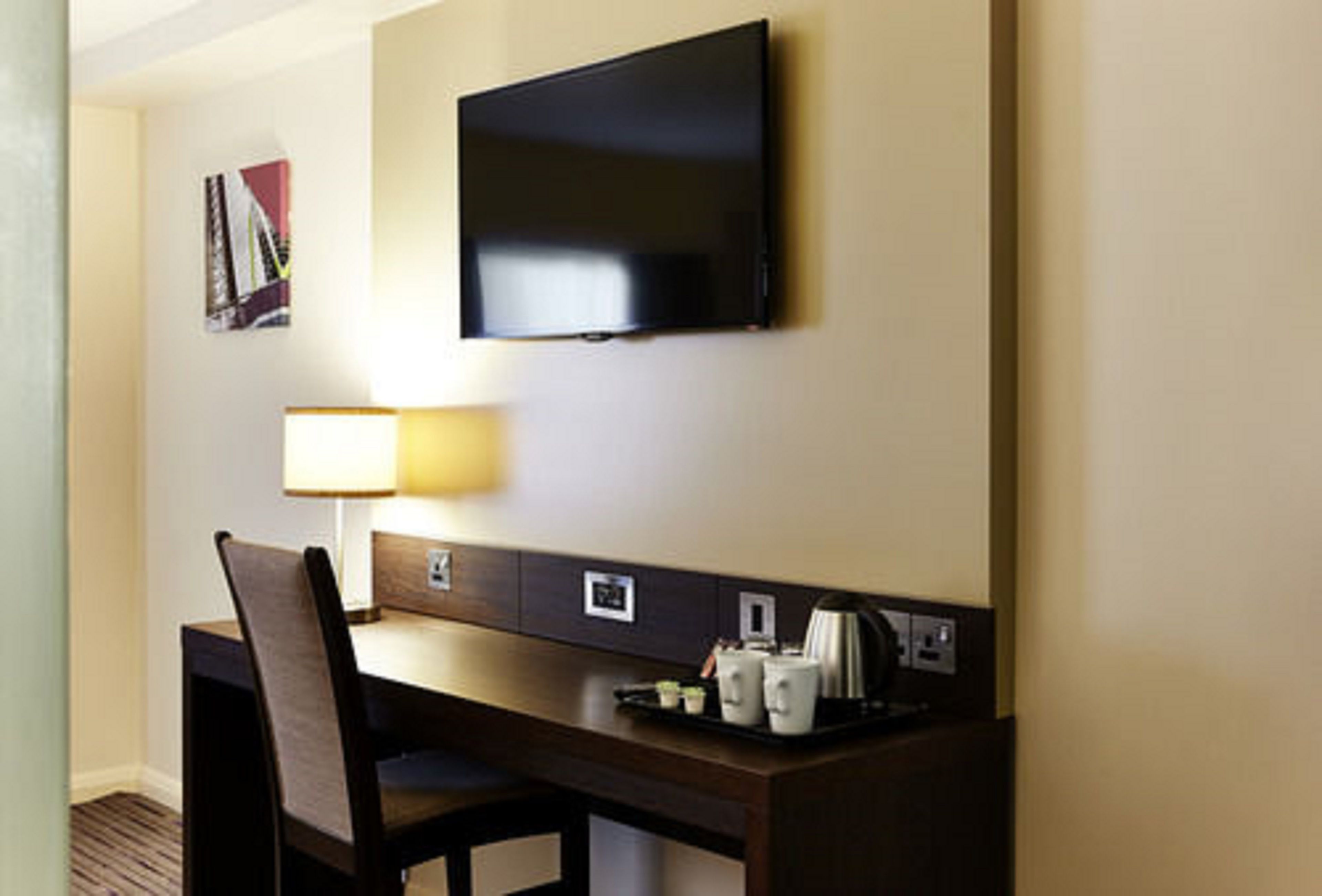 Een hotelkamer met een flatscreen televisie aan de muur, een bureau met een stoel, een lamp en een dienblad met bekers en een koffiezetapparaat.