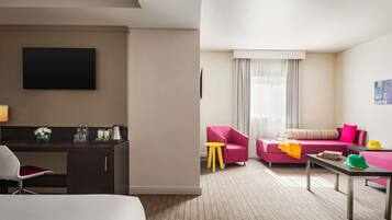 Una habitación de hotel con una cama, un escritorio con silla, un sofá rosa, una mesita y un televisor montado en la pared.