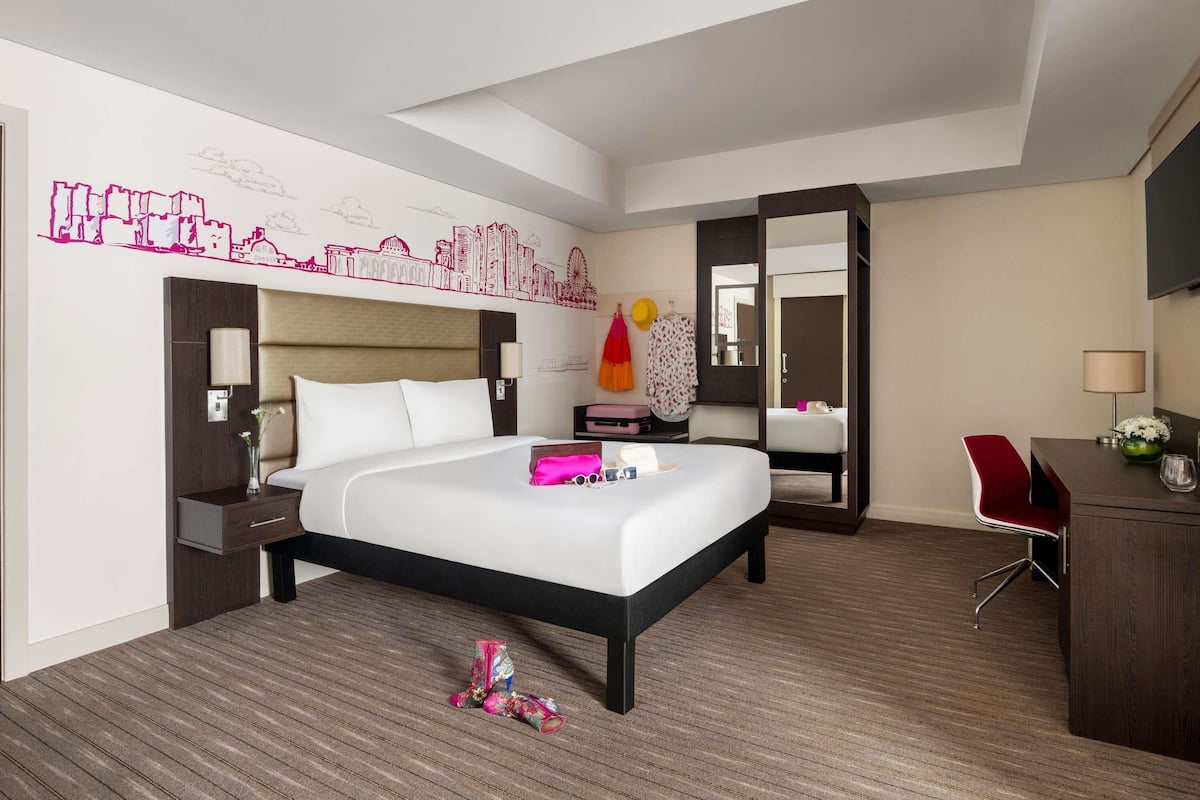 Une chambre d’hôtel avec un grand lit, un bureau, une chaise, un miroir et une décoration murale représentant le panorama de la ville.