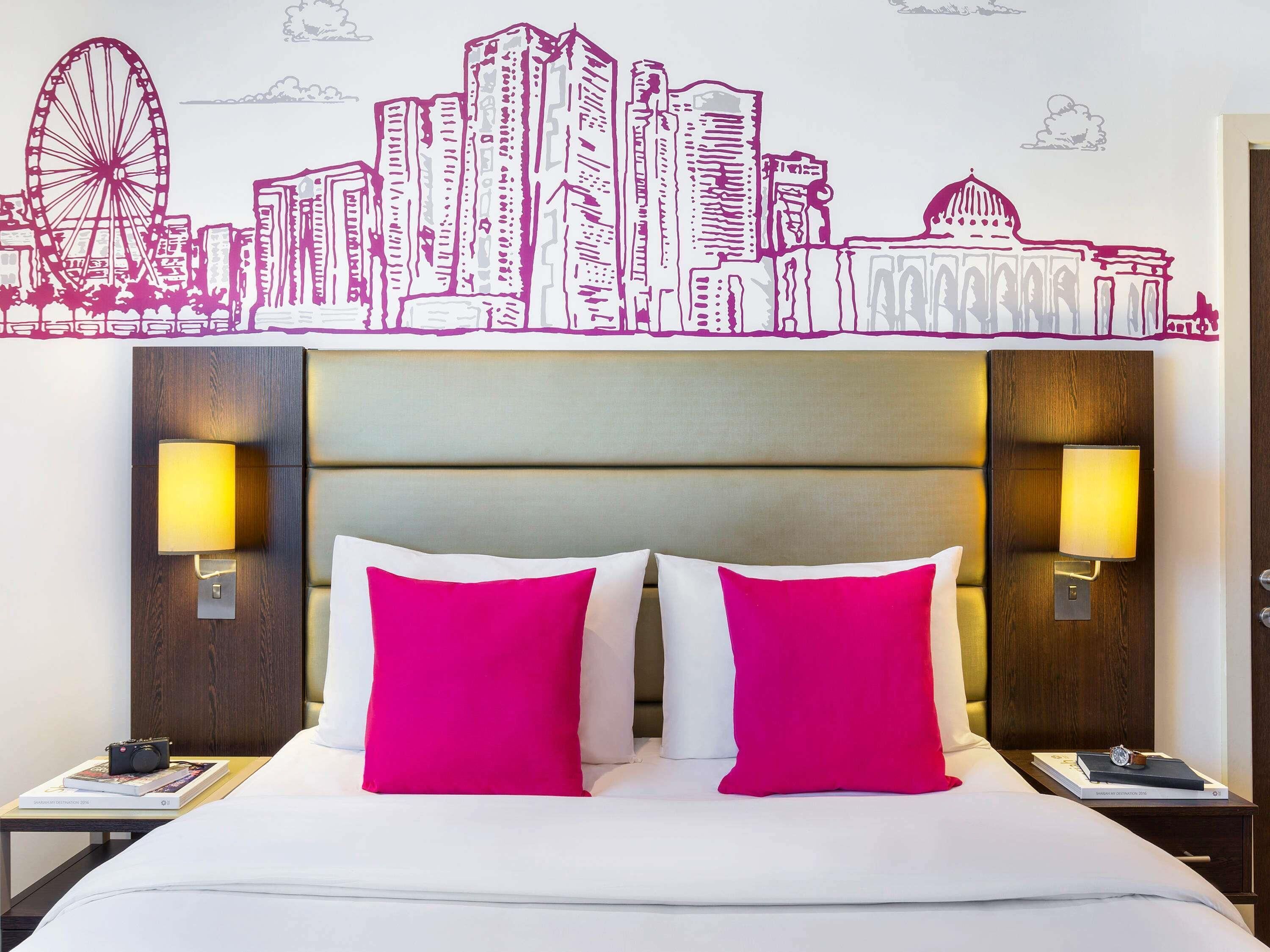 Habitación de hotel con un mural de la ciudad, una cama con ropa de cama blanca y rosa, dos mesitas de noche con lámparas, y una mesita auxiliar con un libro y una cámara.