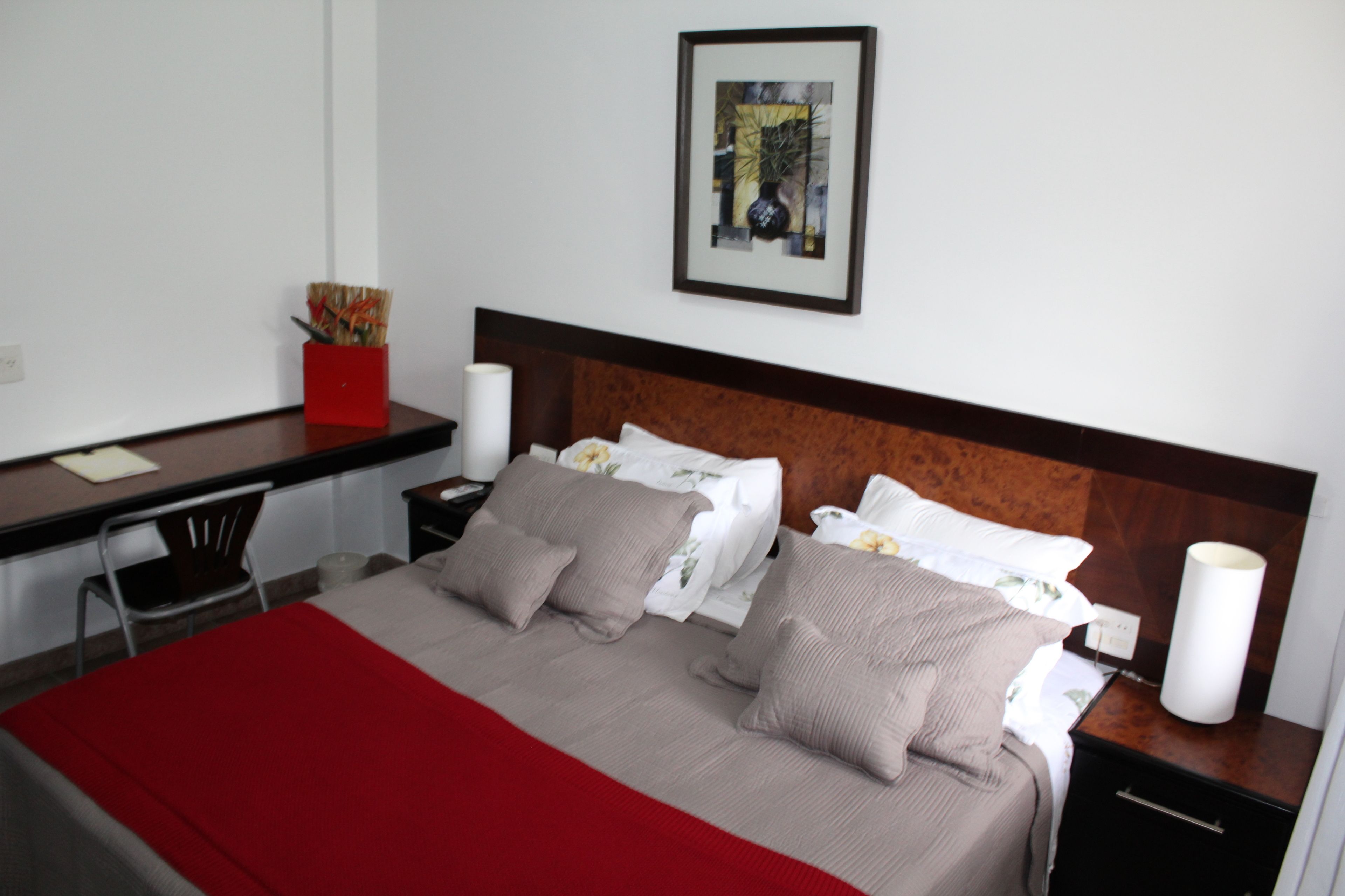 A hotel room with a bed, a desk, a chair, a nightstand, and a framed picture on the wall.