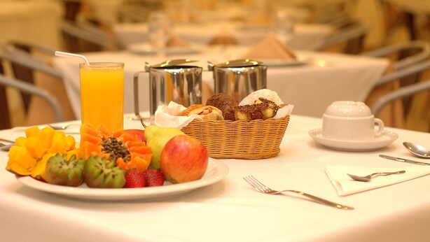 A breakfast setting with a plate of fruit, a glass of orange juice, a basket of pastries, and a cup of coffee on a table. - Hotel Dom Henrique (Timoteo)