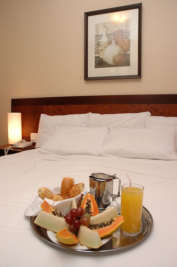 A neatly made bed with white linens, a bedside table with a lamp, a tray with breakfast including bread, cheese, fruit, and juice. - Hotel Dom Henrique (Timoteo)