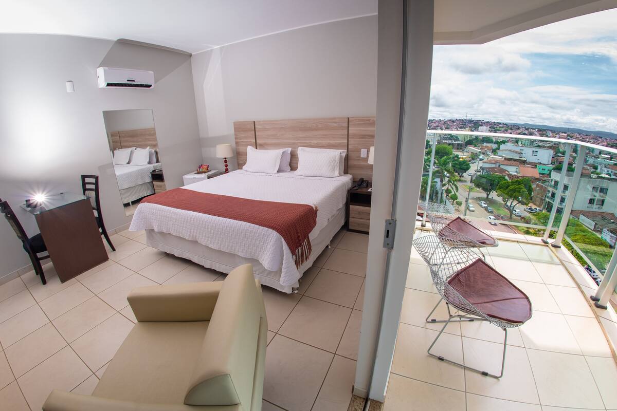 A modern hotel room with a large bed, a balcony with a view of the city, and a seating area with a table.