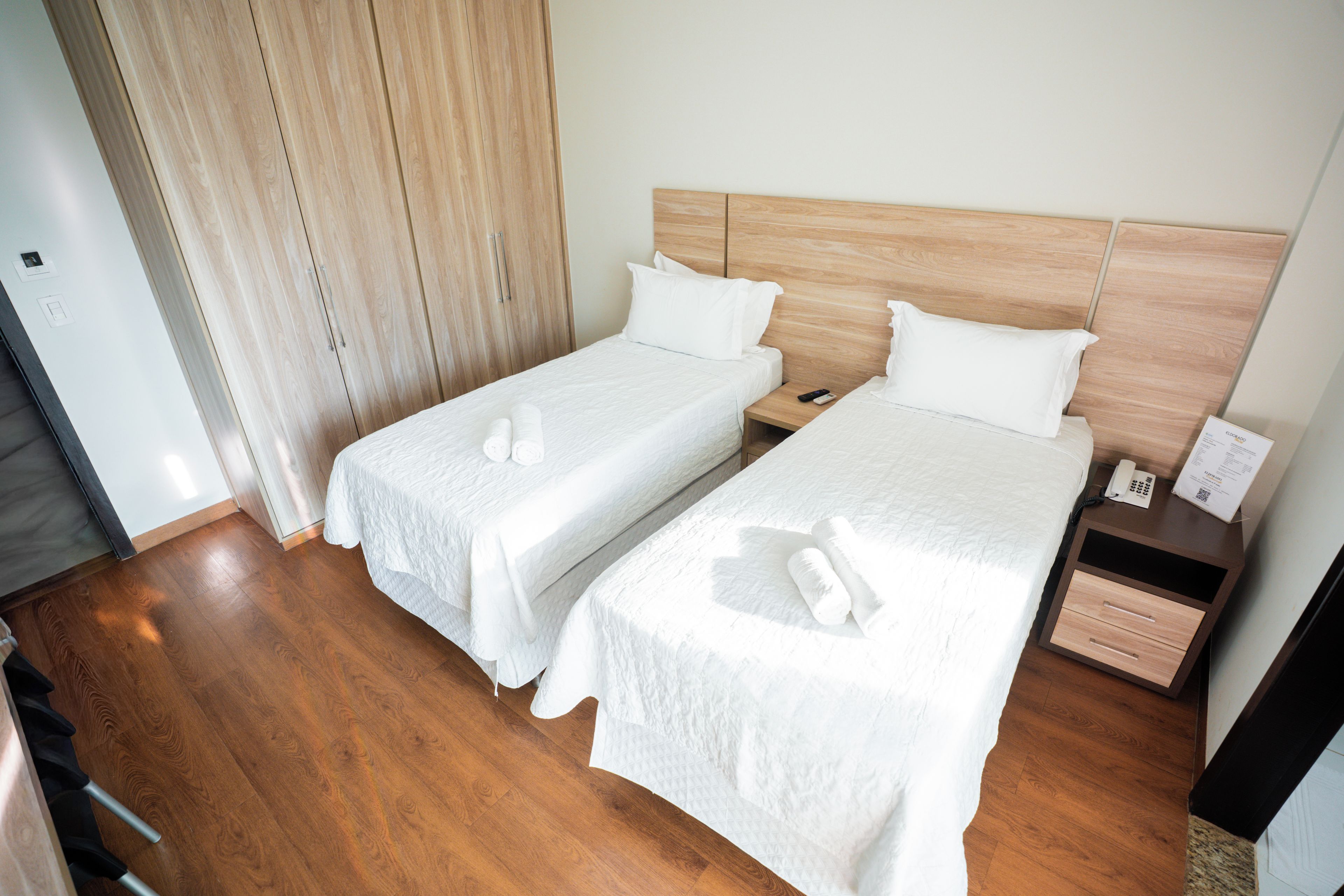 A hotel room with two single beds, wooden headboards, a nightstand with a phone and remote control, and a wooden floor.