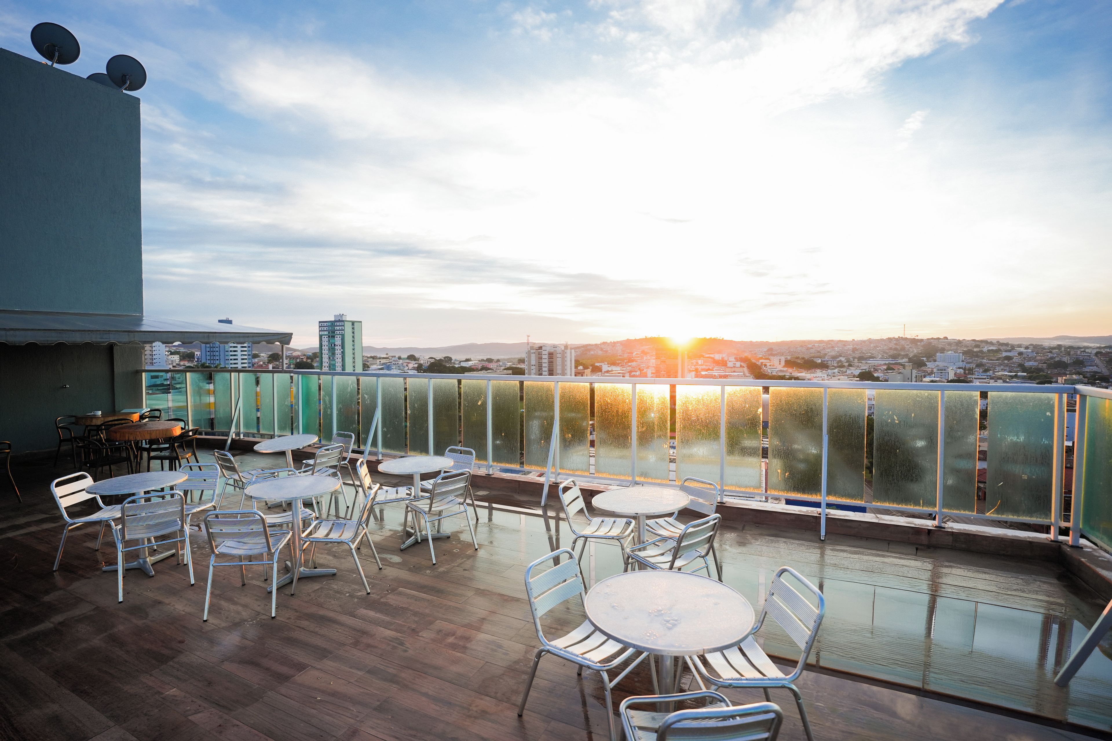 An outdoor terrace with round tables and chairs, overlooking a cityscape during sunset.