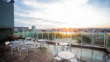 An outdoor terrace with round tables and chairs, overlooking a cityscape during sunset.