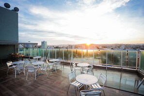 An outdoor terrace with round tables and chairs, overlooking a cityscape during sunset.
