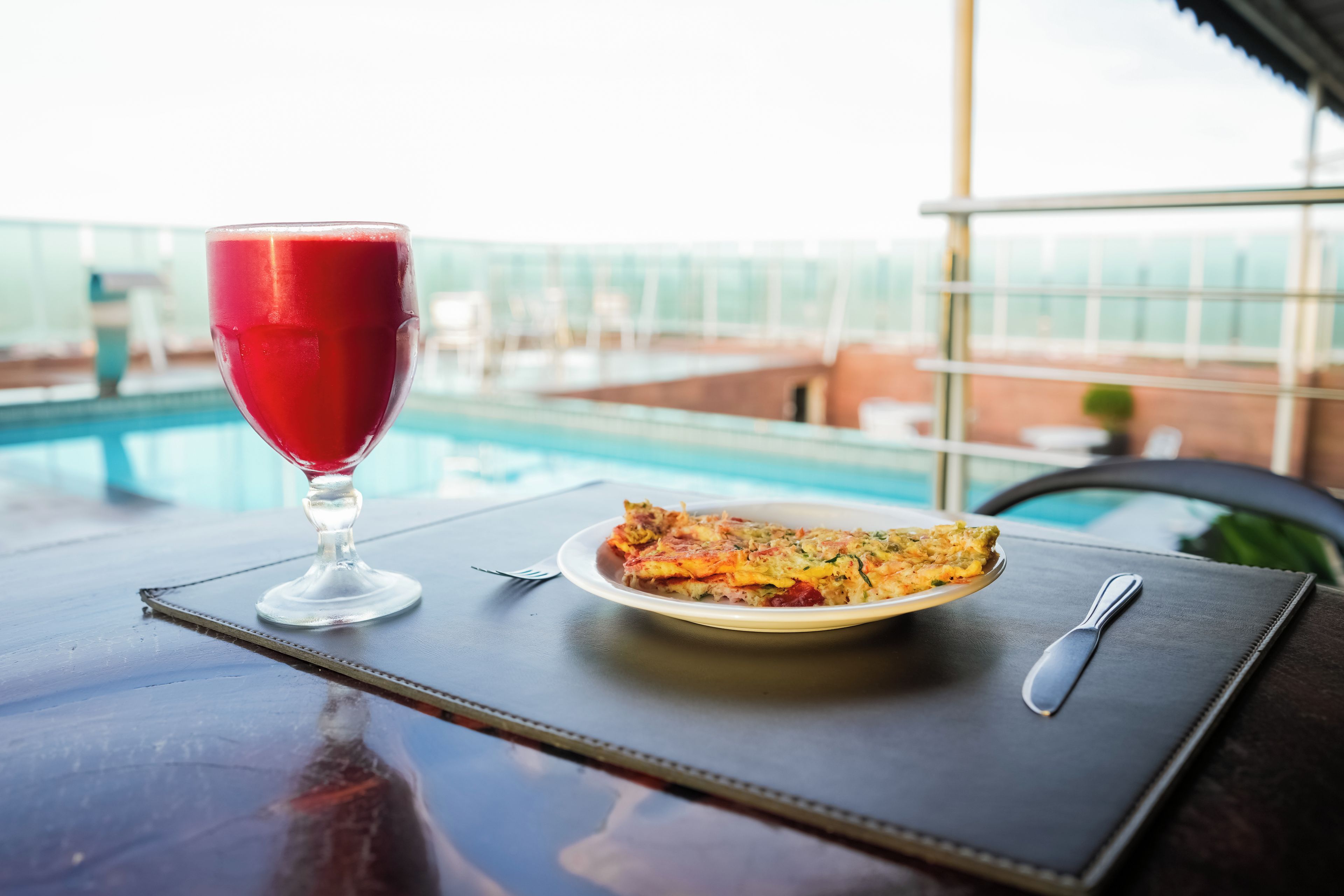 Une table à manger avec un verre d’une boisson rouge, une assiette de nourriture et une vue sur une piscine.