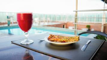 Une table à manger avec un verre d’une boisson rouge, une assiette de nourriture et une vue sur une piscine.
