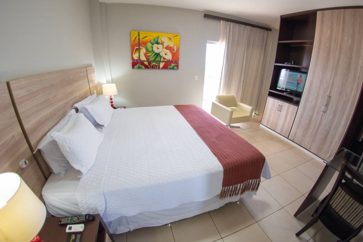 A hotel room with a bed, a TV, a desk with a lamp, a chair, and a wardrobe.