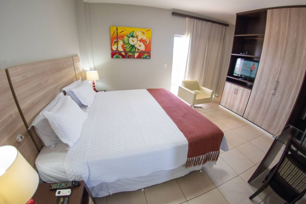 A hotel room with a bed, a TV, a chair, and a painting on the wall.