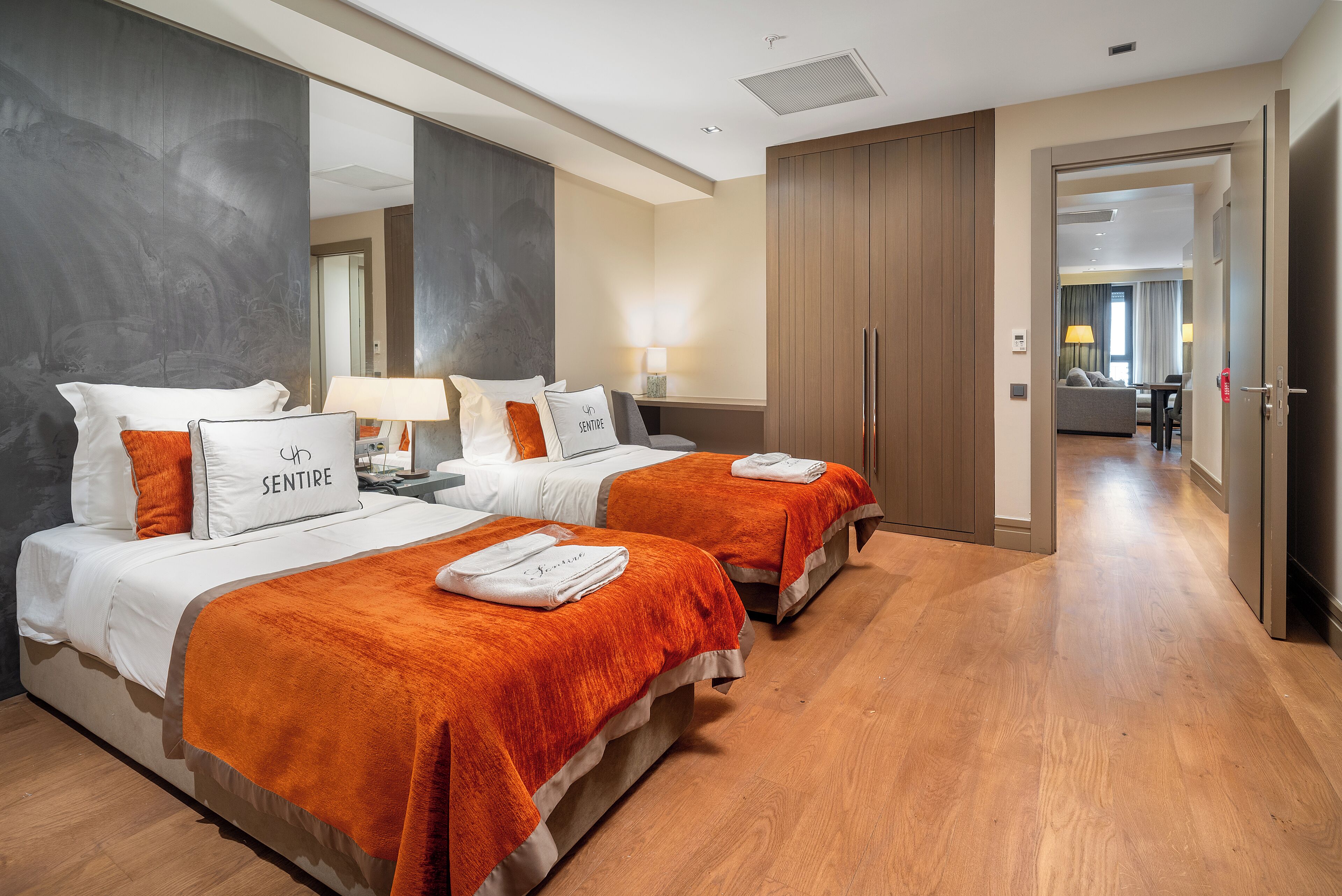 A hotel room with two beds, a large mirror, wooden flooring, and a view into the adjacent room.