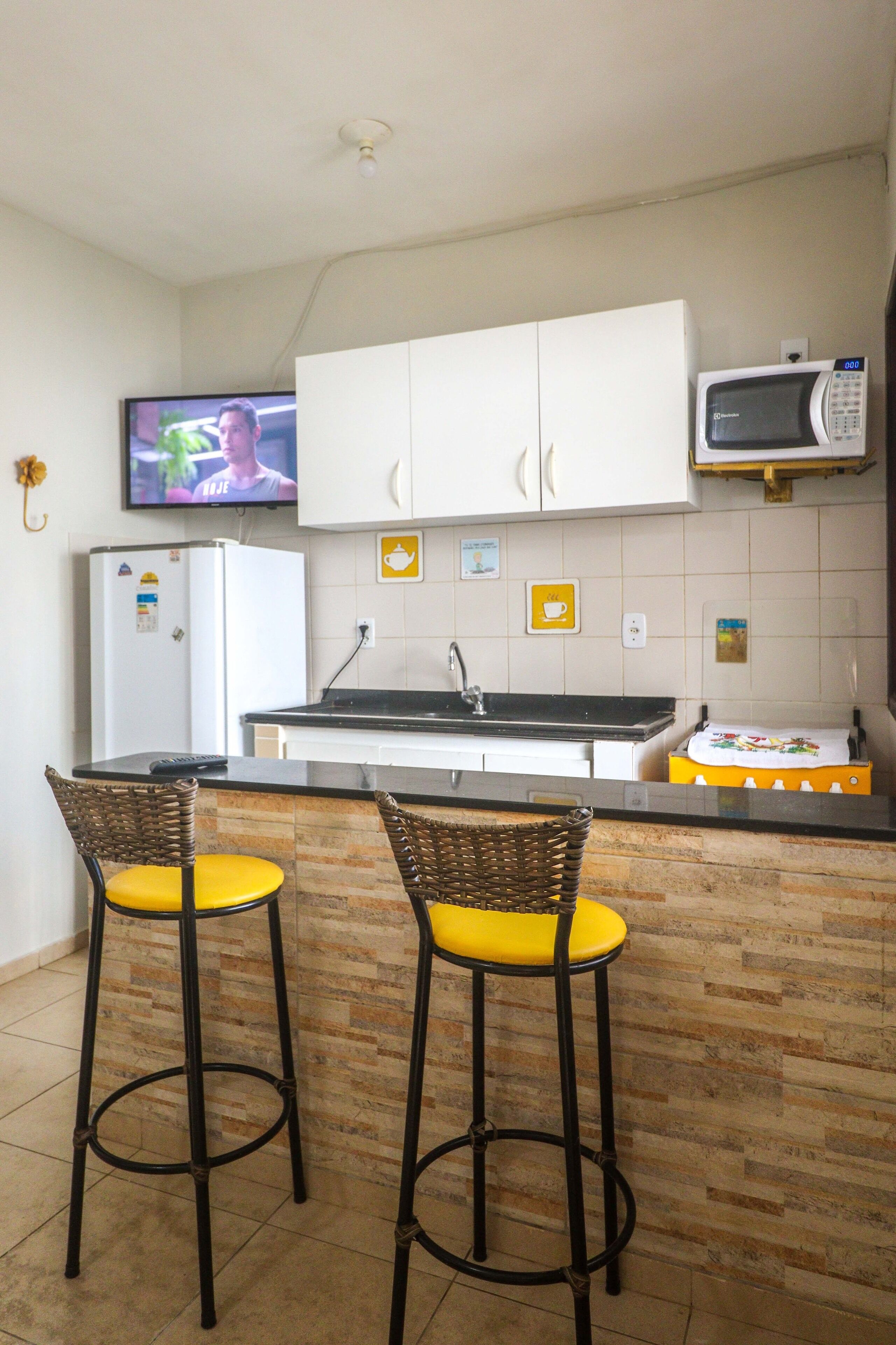 Una cocina compacta con armarios blancos, un microondas, un refrigerador y dos taburetes de bar con cojines amarillos.