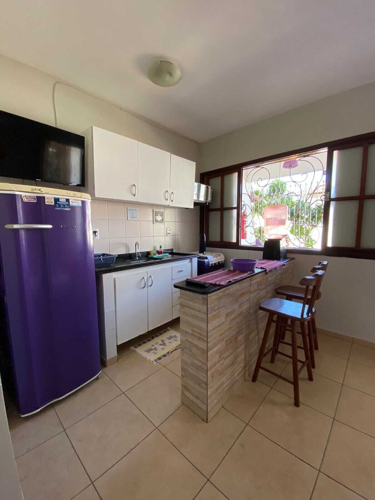 Una cocina con un refrigerador púrpura, gabinetes blancos, piso de baldosas y una ventana con vista a la vegetación.