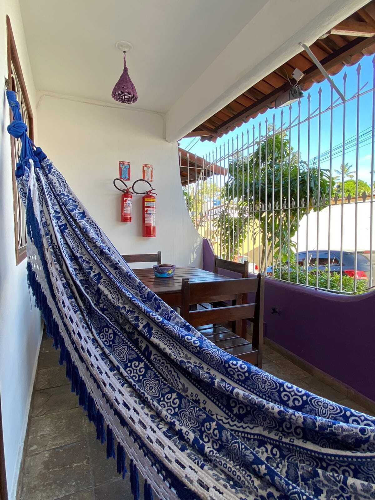 A hammock on a balcony with a table and chairs, fire extinguishers on the wall, and a purple fence.