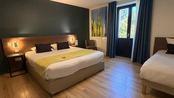 A hotel room with a large bed, a smaller bed, wooden flooring, a nightstand with a lamp, a painting on the wall, and a window with curtains.