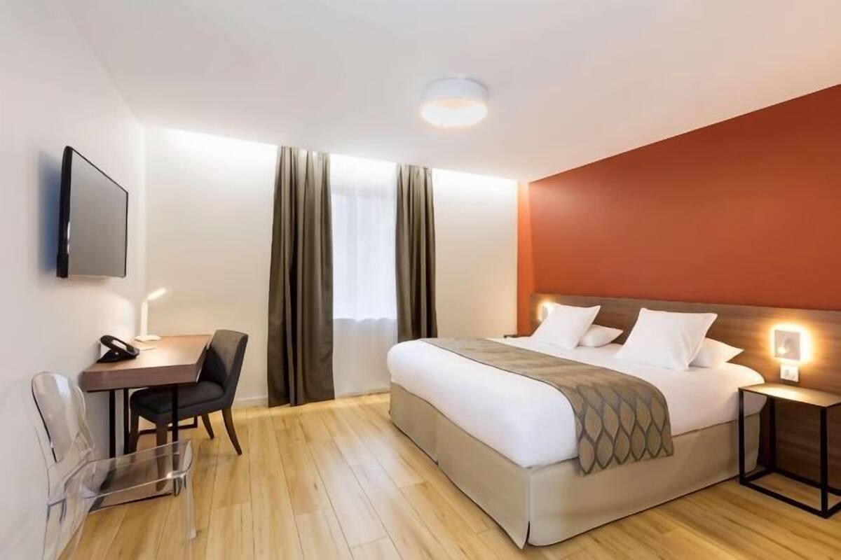 A hotel room with a bed, a desk, a chair, a TV, and a bedside table.