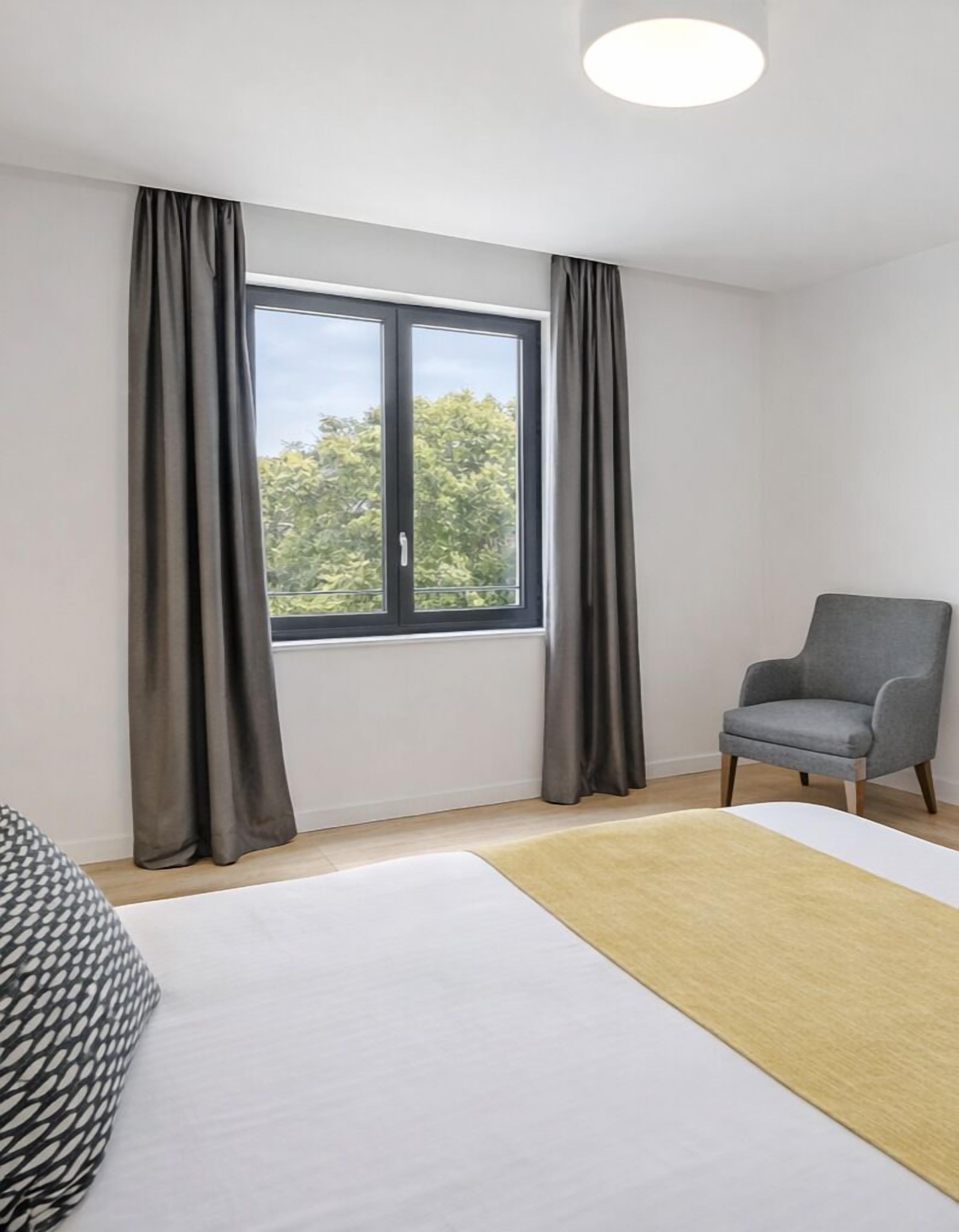 Une chambre moderne avec une grande fenêtre, un fauteuil gris, un lit à motifs et un tapis de couleur claire.
