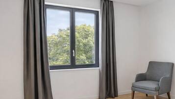 Une chambre moderne avec une grande fenêtre, un fauteuil gris, un lit à motifs et un tapis de couleur claire.