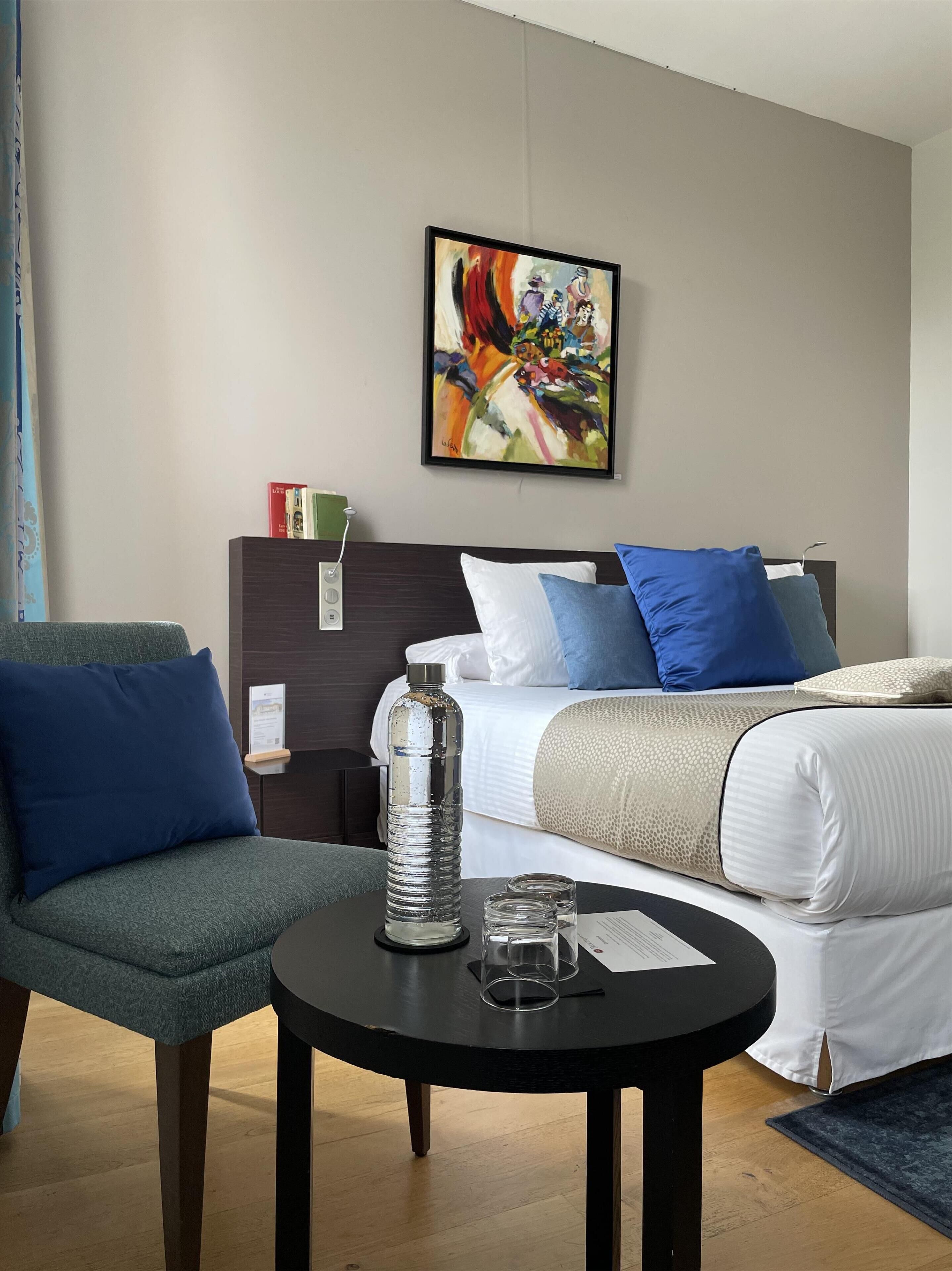 A hotel room with a bed, a chair, a table with a water bottle and glasses, and a painting on the wall.