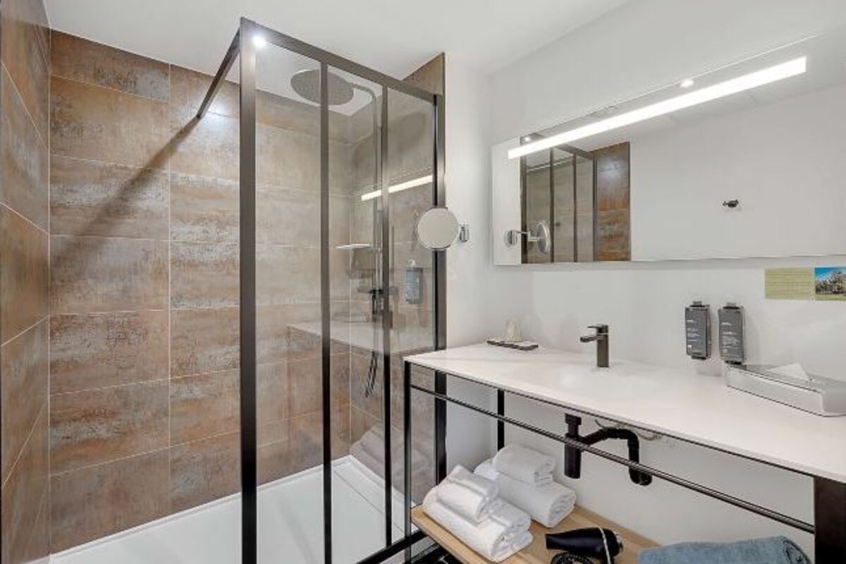 Un baño moderno con ducha envuelta en vidrio, un lavabo blanco con espejo y toallas ordenadas en un estante.
