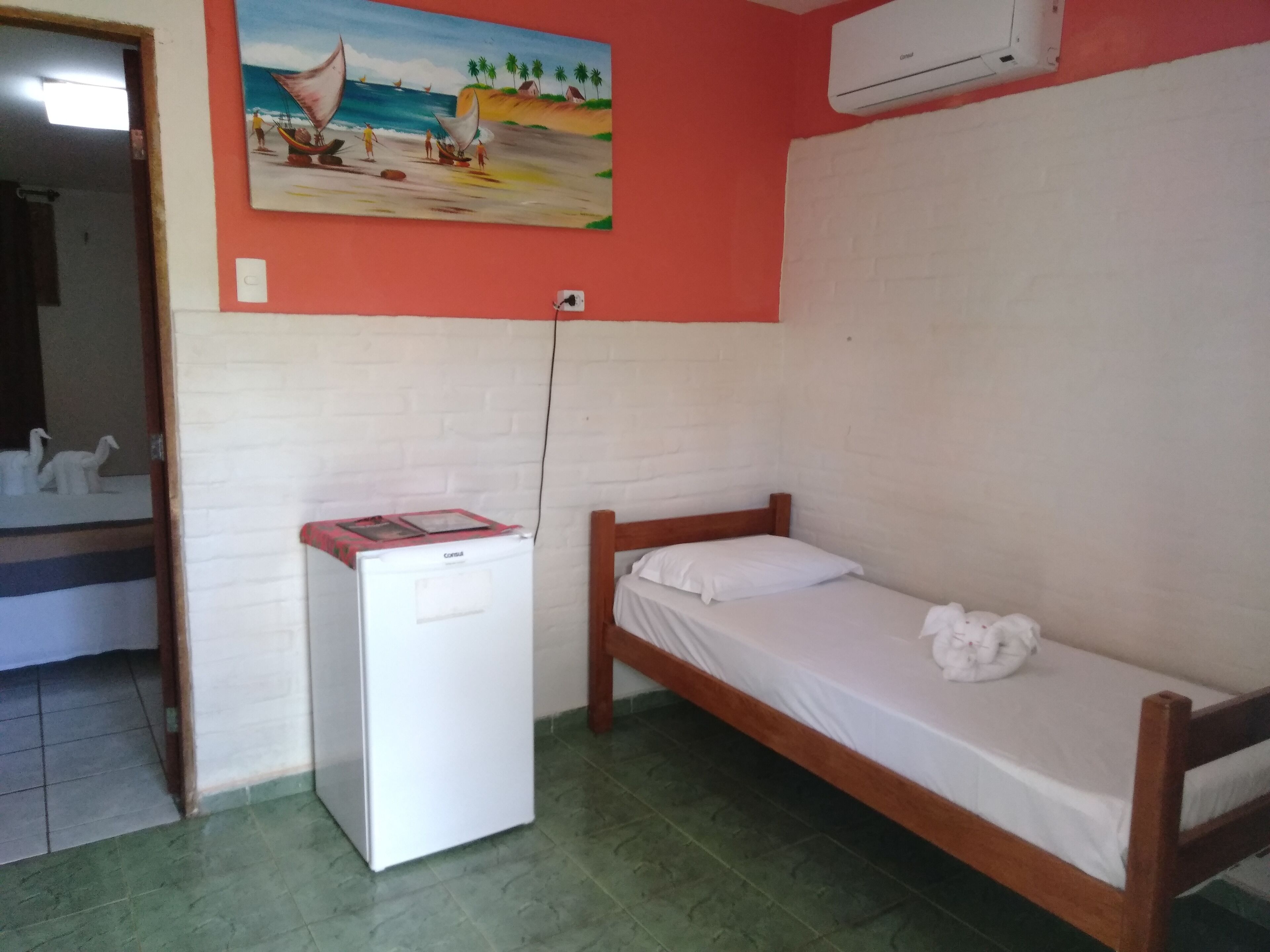 Una habitación pequeña con una cama individual, un refrigerador, un cuadro en la pared y piso de baldosas.
