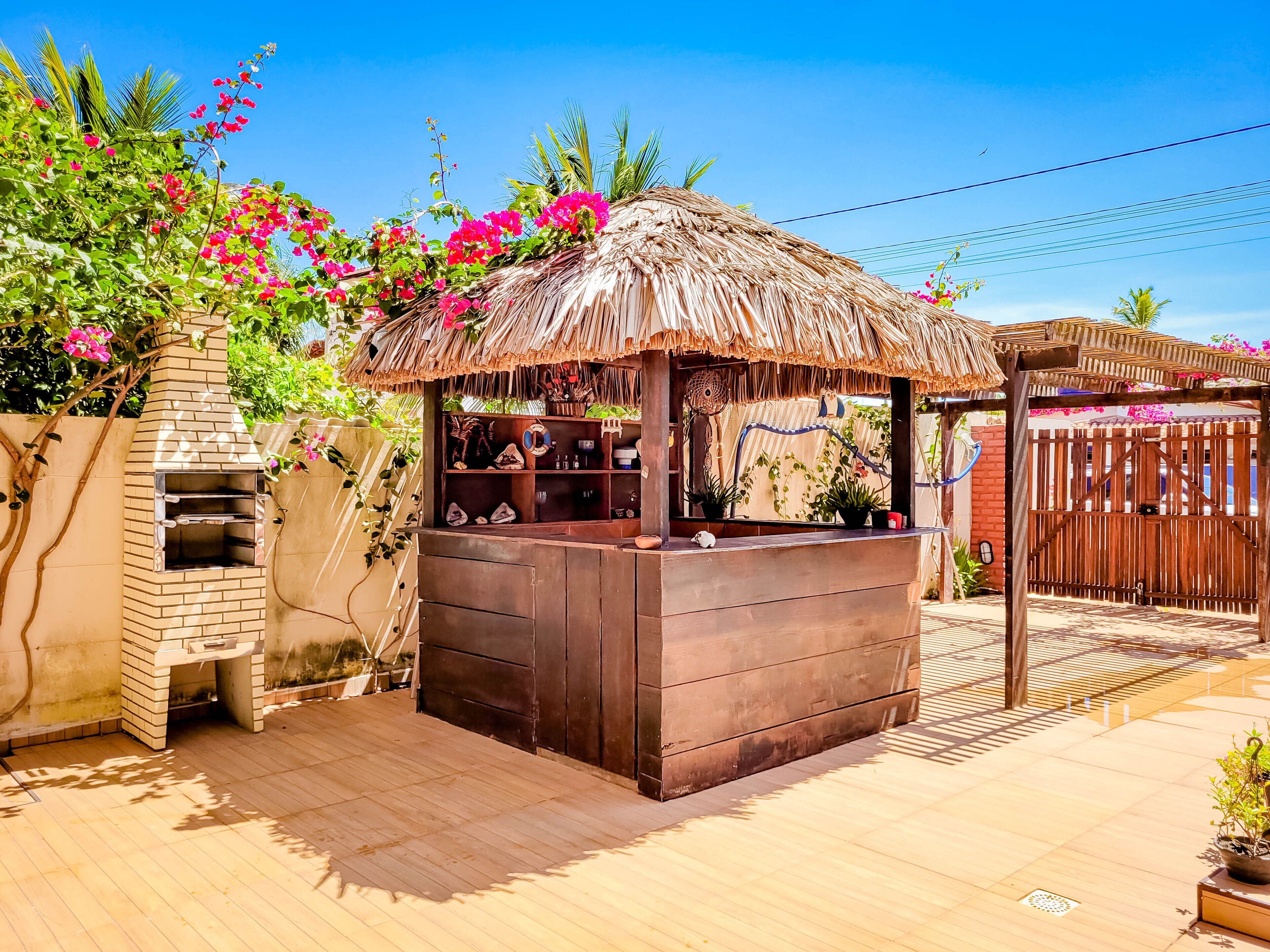 A wooden outdoor bar with a thatched roof, surrounded by lush greenery and pink flowers.