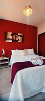 Quarto de hotel com uma cama, parede de destaque vermelha, luminária pendente moderna e mesa de cabeceira com abajur.