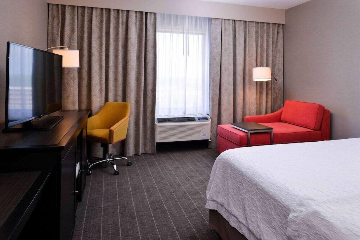A hotel room with a bed, a desk with a TV, a yellow chair, a red sofa, and a window with curtains.