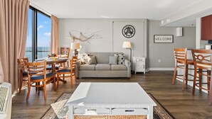 A modern living room with a sofa, dining table, chairs, a white coffee table, and a view of the ocean. - Units at Patricia Grand Resort by Elliott Beach Rentals (Myrtle Beach)