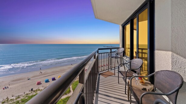 A balcony with a view of the beach and ocean, wicker furniture, and a table with a glass. - Units at Patricia Grand Resort by Elliott Beach Rentals (Myrtle Beach)