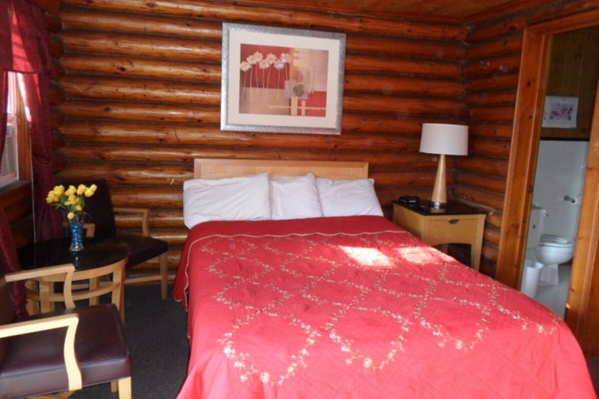 A log cabin room with a bed, a bedside table, a chair, a lamp, and a framed picture on the wall.
