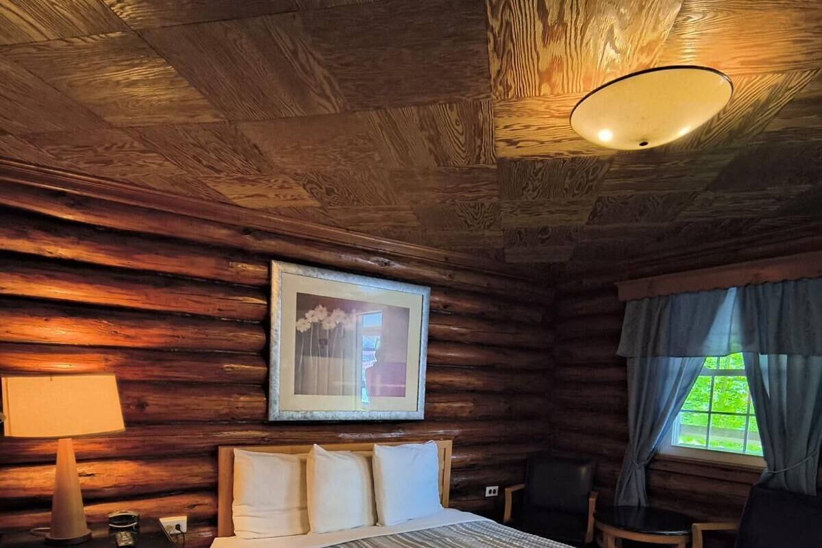 A cozy log cabin room with a large bed, a nightstand with a lamp, a framed picture, and a window with curtains.