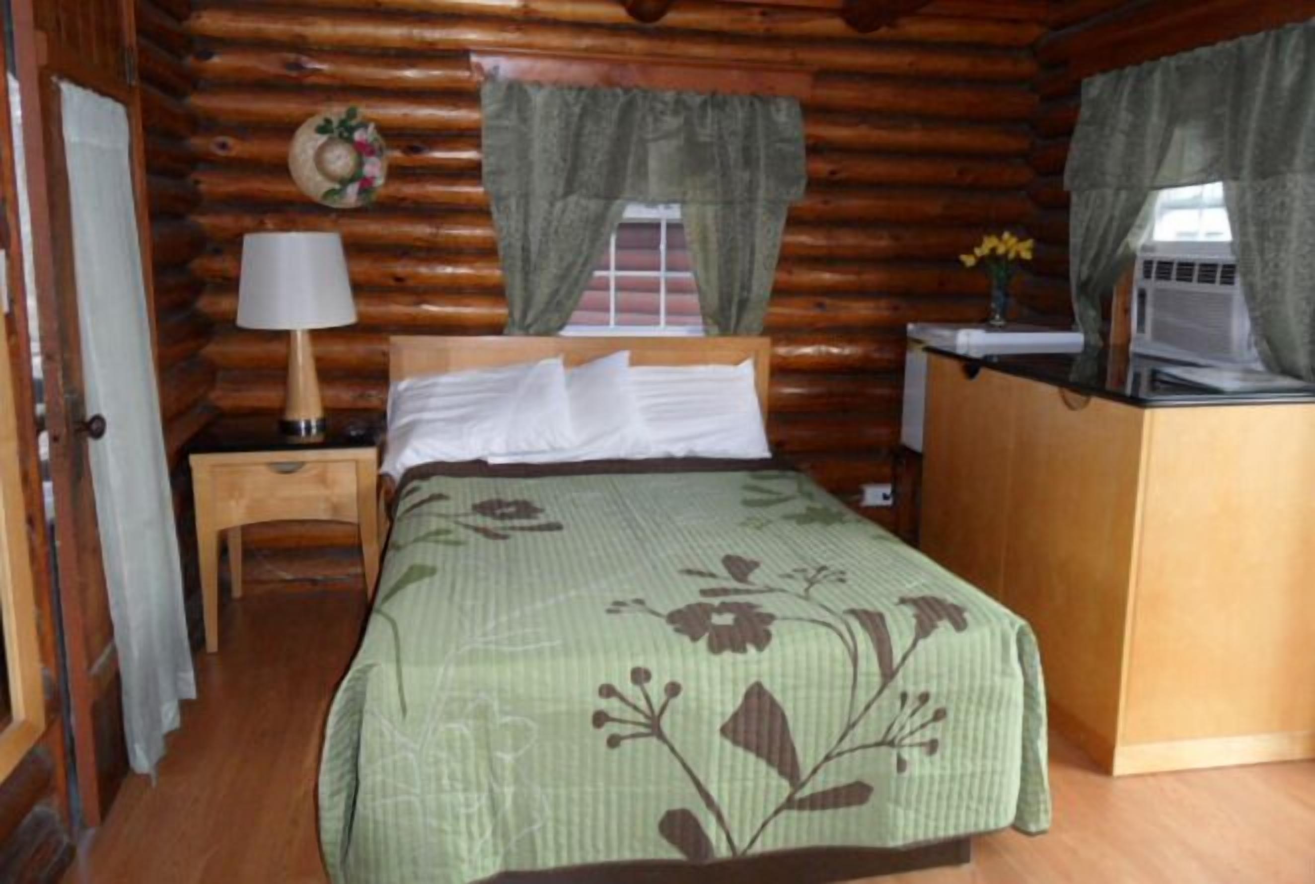 A log cabin room with a bed, a nightstand, a lamp, a desk with a laptop, and a window with curtains.