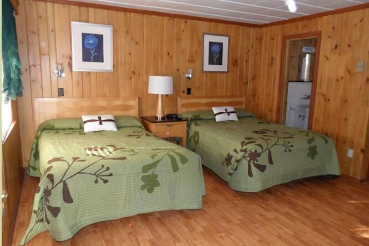 A room with two beds, wooden walls, a nightstand with a lamp, and a refrigerator.