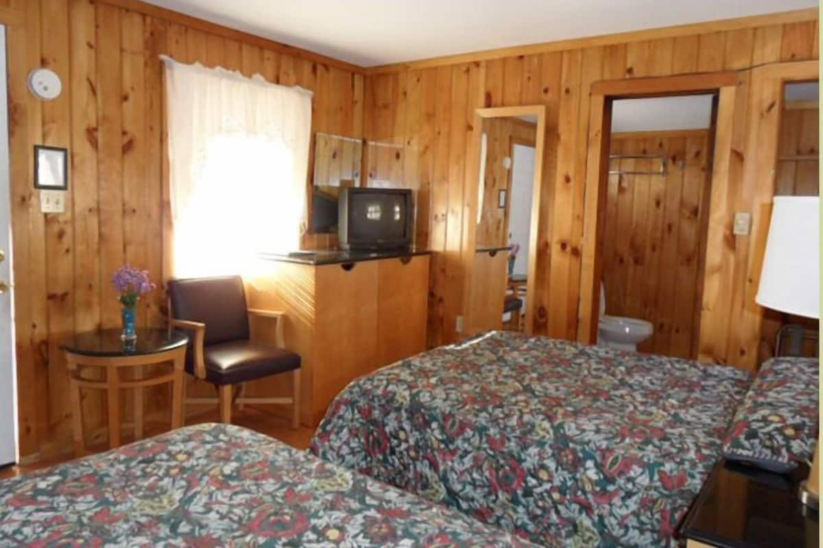 A room with wooden panel walls, a bed with floral bedding, a television on a stand, a chair, and a small table with a vase.