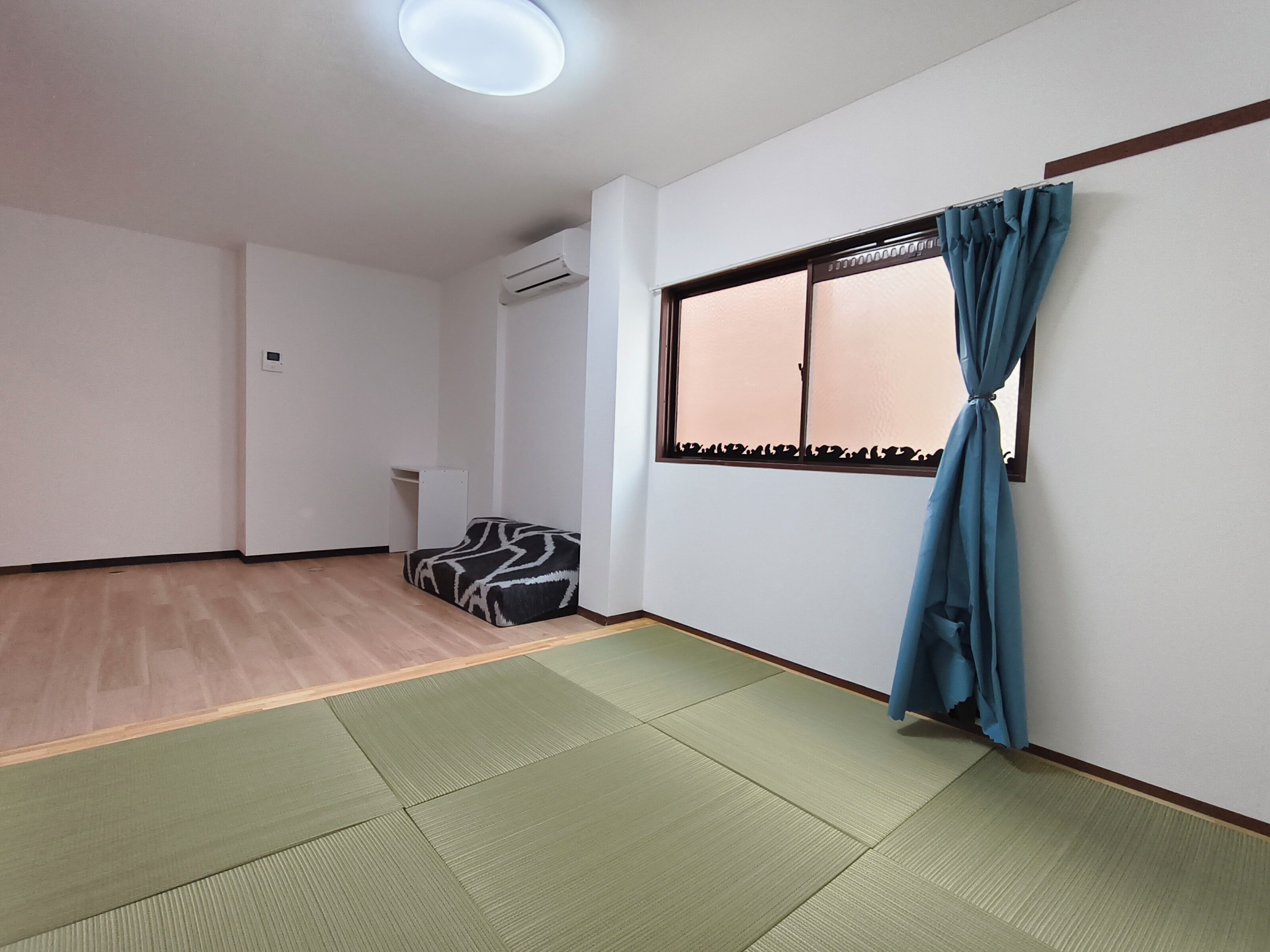Ein Zimmer mit Tatami-Matten, einem Vorhangfenster, einer Klimaanlage und einer kleinen Sitzecke.