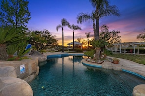 A swimming pool with surrounding rocks and palm trees, a gazebo, and a sunset in the background. - 5-acre Luxe 5B Getaway @Murrieta | Pool, Hot Tub (Murrieta)