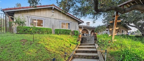 A house with a stone facade, a wooden staircase leading to a covered porch, and a garden with a variety of plants.