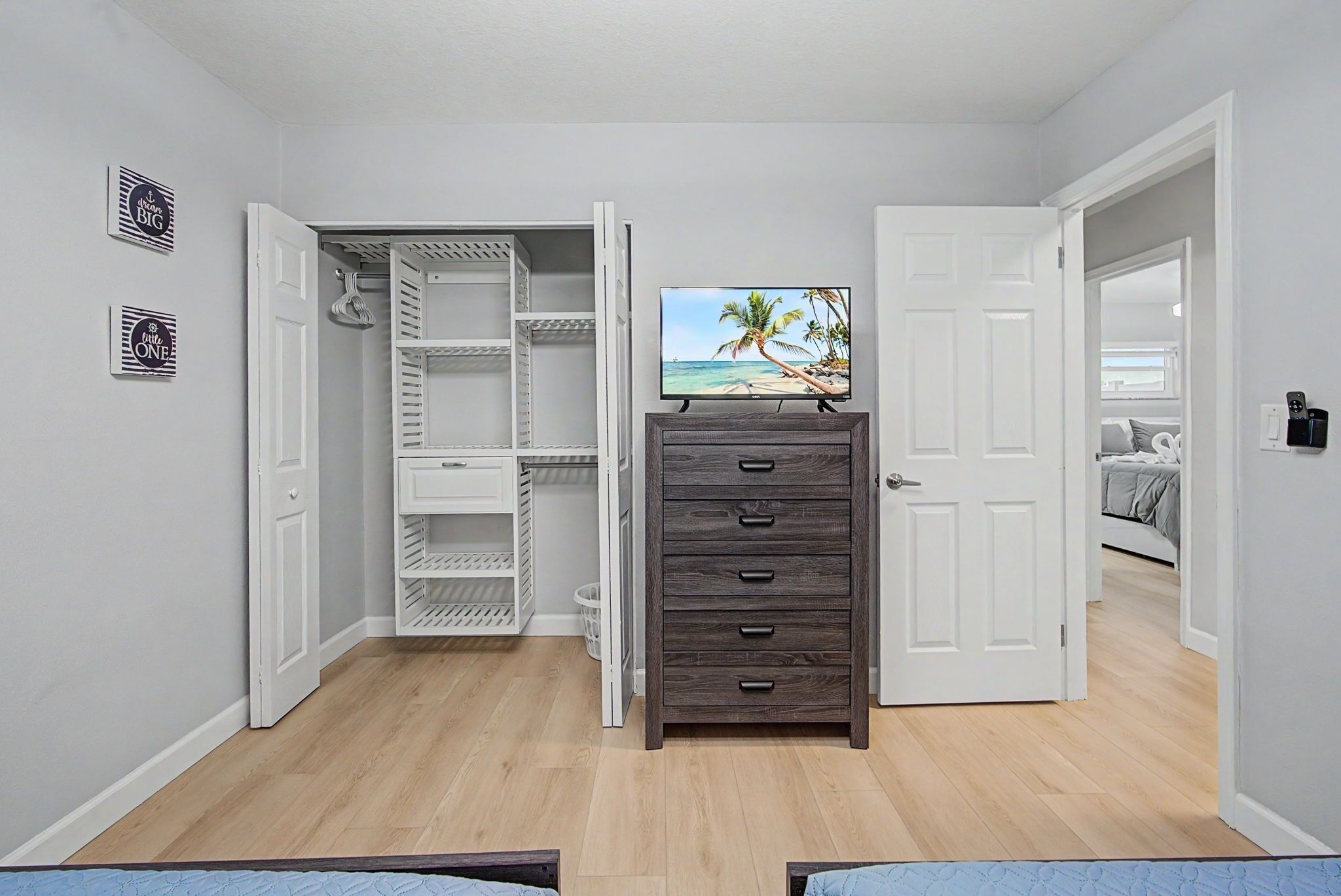 Una habitación con un aparador de madera, un televisor sobre un mueble bajo, un armario blanco con las puertas abiertas y una cama al fondo.