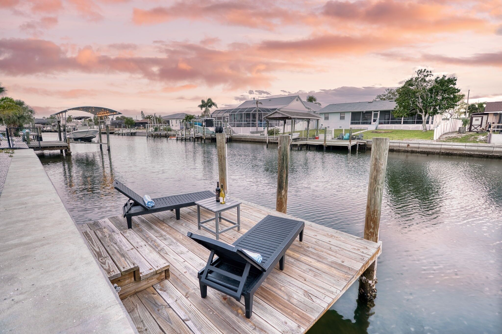A waterfront dock with lounge chairs and a table, a small boat docked, and houses in the background.