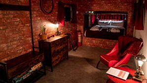 A room with a brick wall, a bed with red curtains, a red chair, a desk with drawers, and a small table with a lamp. - Cabin Sleeps 2 With Pets Welcome (Skegness)