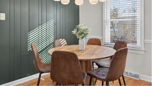 Un coin repas avec une table ronde en bois, six chaises marron, un vase blanc orné de fleurs et un lustre moderne.