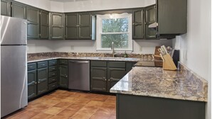Une cuisine avec des meubles de couleur vert foncé, un réfrigérateur en acier inoxydable, un plan de travail en granit et une fenêtre donnant sur les arbres.