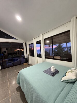 A bedroom with a bed, a view of a pool and outdoor area, and a sunset view through the windows.