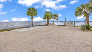 A sandy beach with palm trees, a concrete walkway, and a wooden pier extending into the water. - Cozy Melbourne Retreat | Eau Gallie Arts | Wi-Fi (Melbourne)