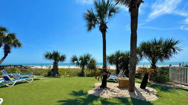 A beach view with palm trees, a grassy area with lounge chairs, and a wooden deck. - Mariner’s Escape - Ocean View 3BR Sleeps 12 (Myrtle Beach)