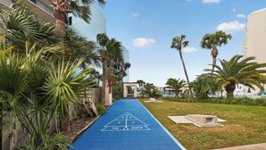 A blue shuffleboard court with a triangular target, surrounded by palm trees and a building. - Beachfront Studio - Ocean View • Pool • Sleeps 4 (Panama City Beach)